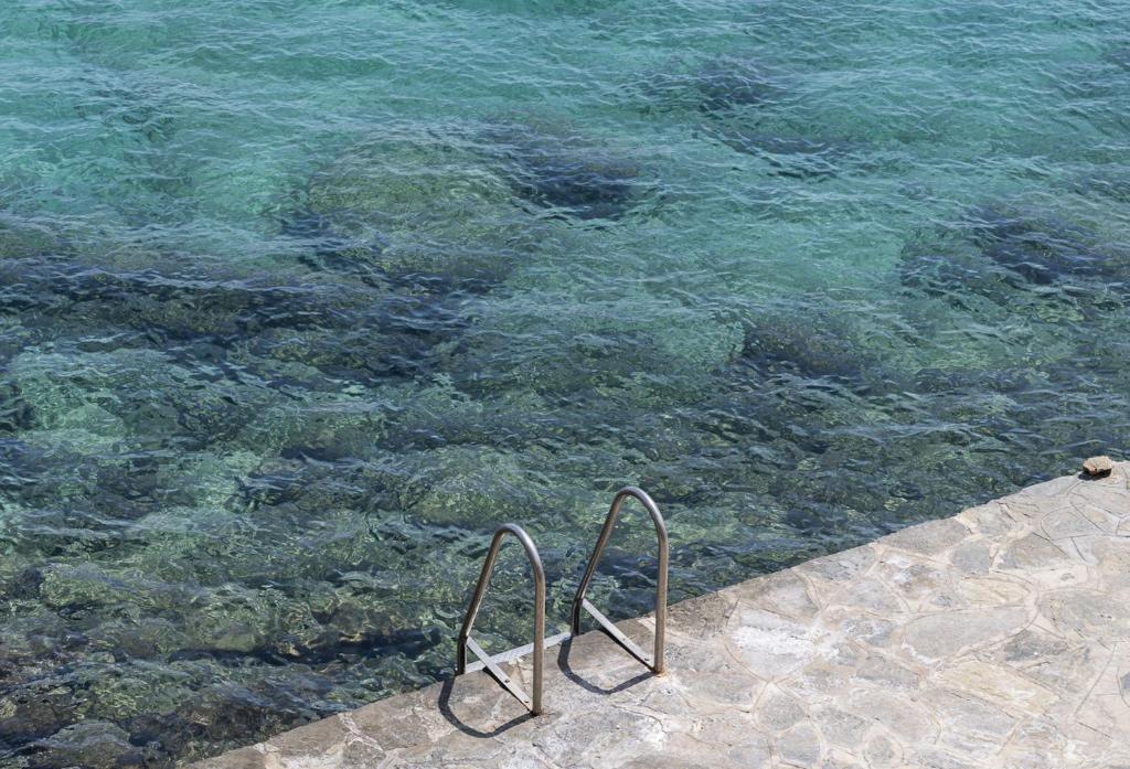 Skats uz skaidru ūdeni ar akmeņiem apakšā, blakus metalizēta kāpņu margas. Ideāla vieta peldēšanai un atpūtai pie jūras.