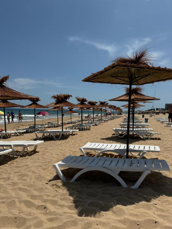 Pludmales ainava ar baltām sauļošanās krēsliem un salmiem, kas sniedz ēnu, un jūras viļņiem fona. Ideāli piemērota atpūtai un relaksācijai saulainā dienā.