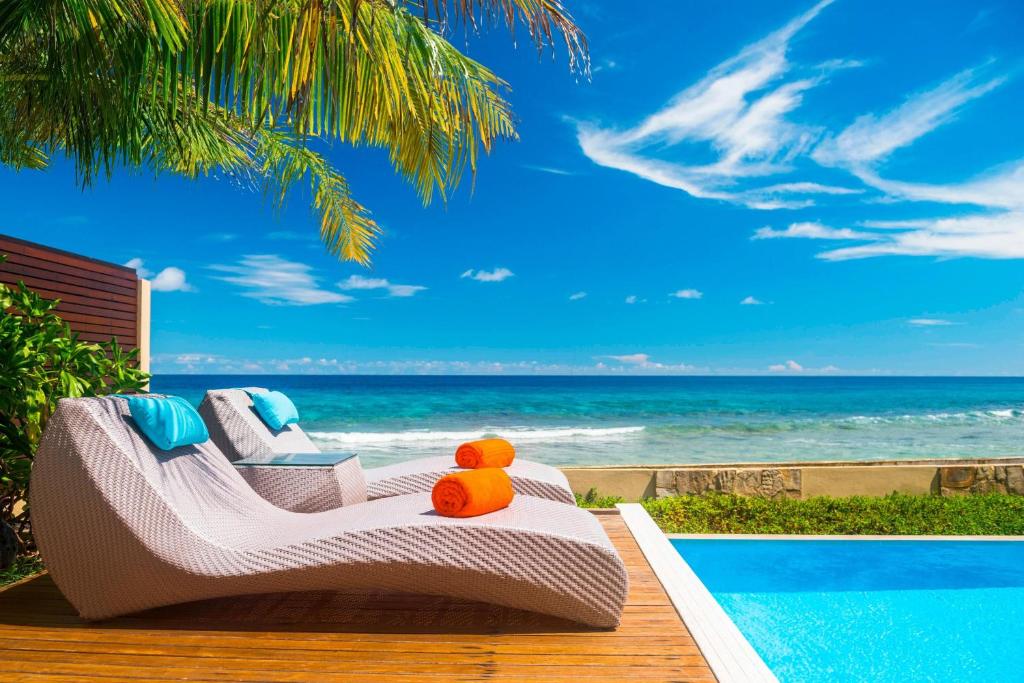 Saulainā pludmales atpūtas zona ar divām modernām atpūtas krēslām un spilveniem, kas novietoti blakus baseinam un jūrai, ar skaidru zilo debesu fonu.