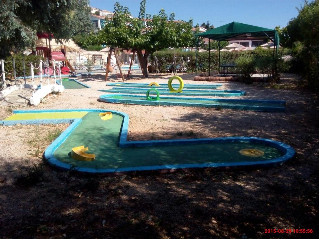 Minigolfa laukums ar zaļām un zilām takām, kas izvietots saulainā vidē ar kokiem un šūpolēm. Ideāla vieta atpūtai un izklaidei.