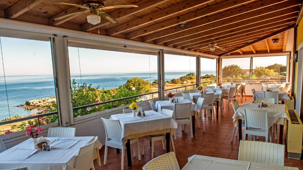 Gaiša restorāna telpa ar skatu uz jūru, kur galdi ir sakārtoti ar baltām galdautām un ziediem. Svaigs, mierīgs atmosfēra, ideāla vieta maltītei.