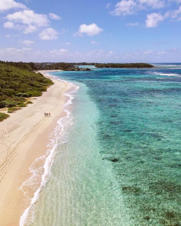 Skaista pludmale ar smilšainu krastu un skaidru ūdeni, kurā redzami koraļļi. Tālā pludmalē pastaigā trīs cilvēki, fonā redzama zaļa veģetācija un zils okeāns.