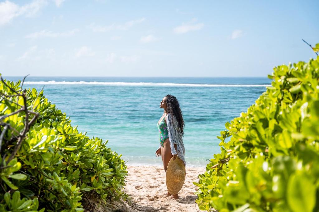 Sieviete pludmalē, tērpusies peldkostīmā un ar platmali rokās, stāv pie ūdens, apņemta ar zaļām krūmiem. Aiz viņas redzams gaiši zils okeāns un skaidras debesis.