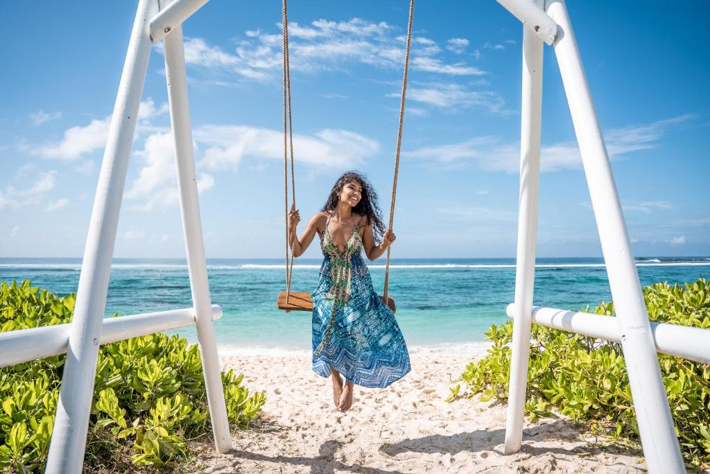 Sieviete ar garām, viļņainām matiem, tērpusies krāsainā kleitā, šūpojas uz šūpolēm pludmalē ar zilu jūru un debesīm fonā.