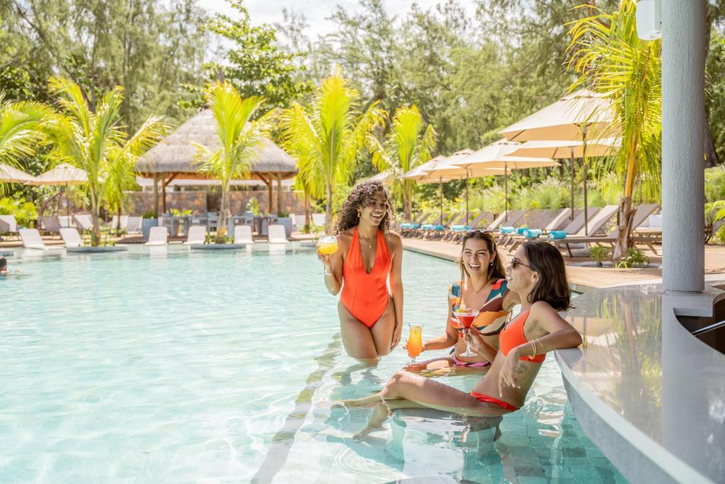 Trīs draudzenes izbauda atpūtu pie baseina, tērpušās peldkostīmos un turēdamos kokteiļus. Apkārt redzami palmām un sauļošanās krēsliem.