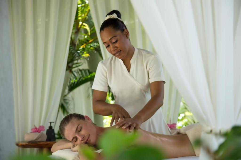 Masāžas sesija tropiskā vidē, kur profesionāla masiere sniedz relaksējošu masāžu apmeklētājam. Jūtu atmosfēra, ko rada zaļie augi un gaisīgas aizkari.