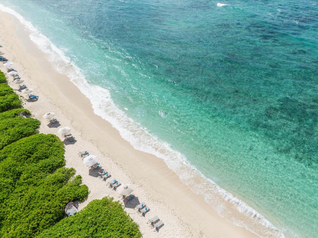 Bāzes pludmale ar smilšu krastu un zilgani zaļu ūdeni, kur redzami atpūtas krēsli un saulessargi. Apkārt pludmalei aug zaļš krūmājs, radot mierīgu atmosfēru.