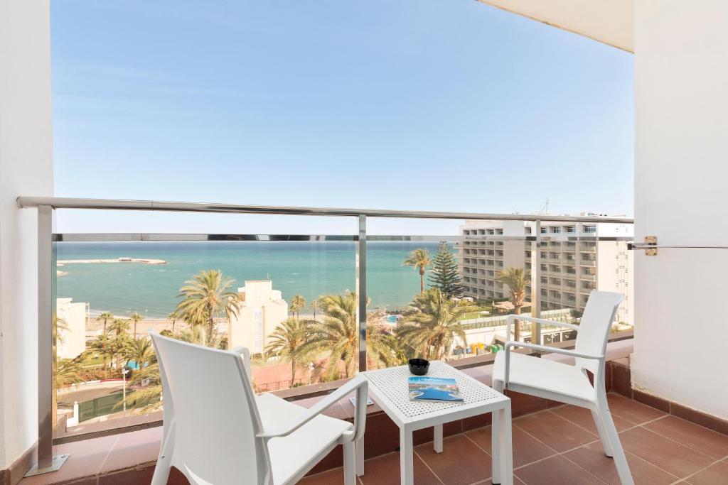 Balkons ar skatu uz jūru un palmām, kurā atrodas divas baltas krēslas un galdiņš. Ideāla vieta relaksācijai un atpūtai.