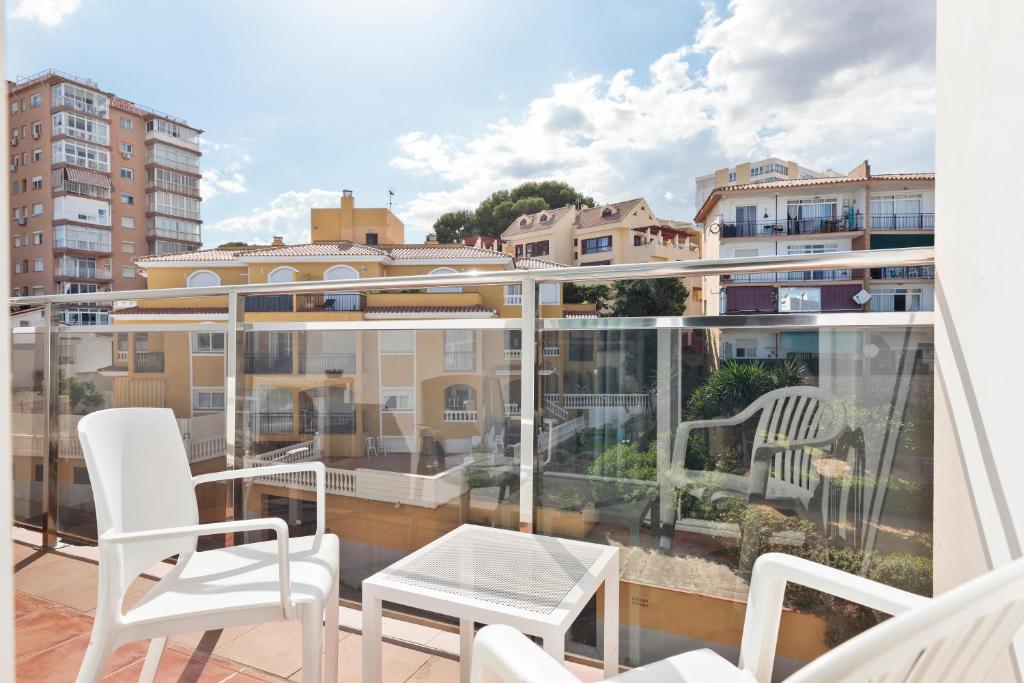 Balkons ar baltām mēbelēm, no kura paveras skats uz blakus esošām ēkām un zaļajiem apstādījumiem. Gaisīgs un saulains laiks ar daļēji mākoņainu debesīm.