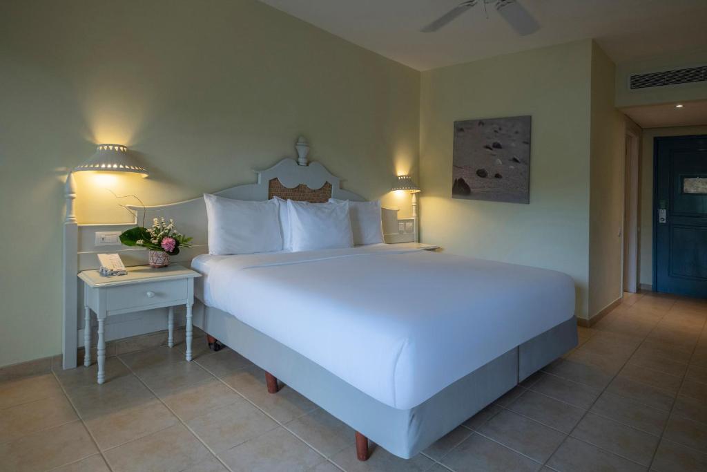 Mūsdienīgs viesnīcas numurs ar plašu gultu, baltām gultasveļām un elegantu naktsgaldiņu. Telpā ir maiga apgaismojums un dekoratīvs attēls uz sienas, radot relaksējošu atmosfēru.