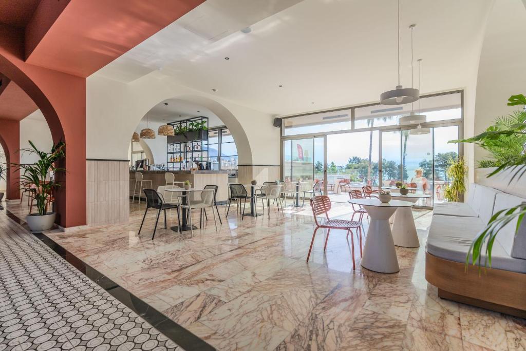 Moderns kafijas veikals ar elegantu interjeru, kas ietver arkas un plašus logus, kas sniedz skatu uz dabu. Telpa ir dekorēta ar augiem un stilīgām mēbelēm, radot patīkamu atmosfēru.