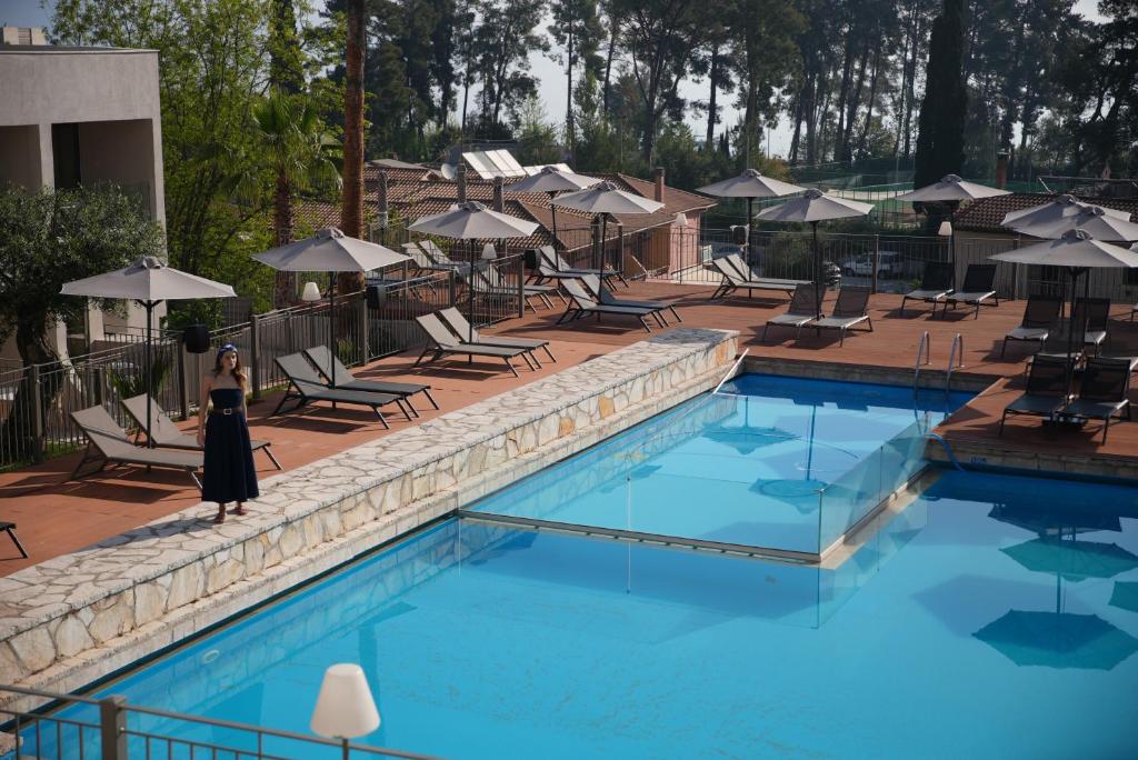 Baseins ar skaidru ūdeni, apkārt atpūtas krēsli un saulessargi, blakus stāv sieviete tumšā kleitā.