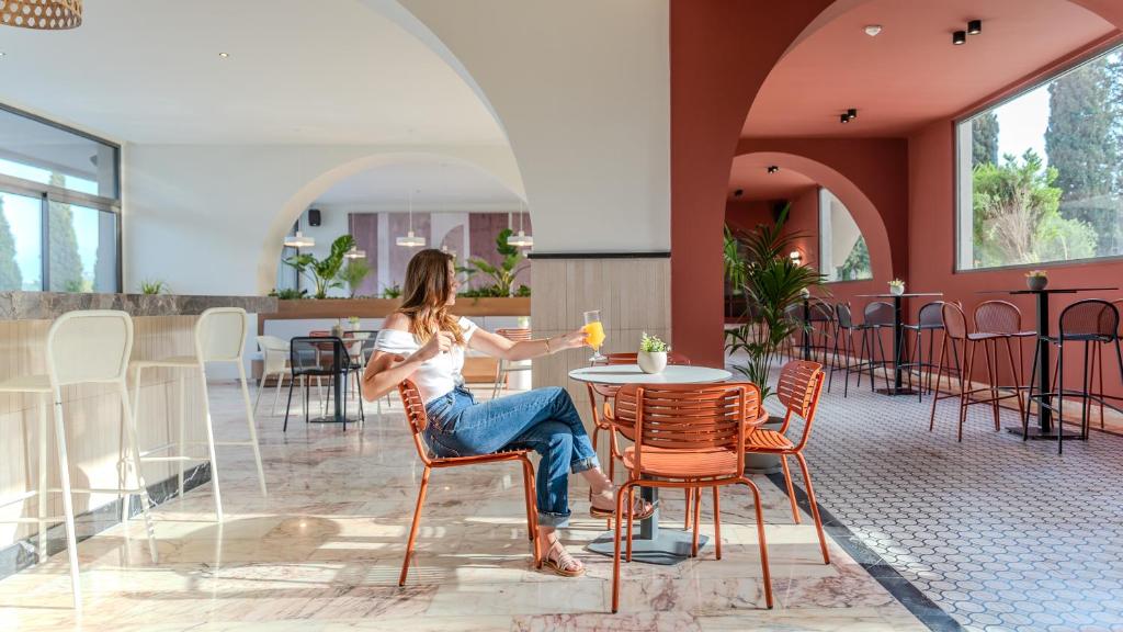 Mūsdienīgs kafijas veikals ar gaišu interjeru, kurā sēž sieviete un bauda dzērienu pie galda, apstādīta ar zaļām augiem un stilīgām mēbelēm.