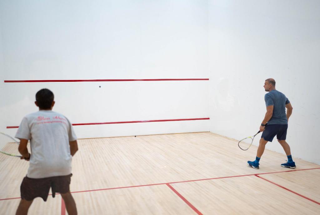 Divi spēlētāji spēlē skvošu sporta zālē, fokusējoties uz bumbu. Sporta zāles sienas ir baltas ar sarkaniem līniju marķējumiem.