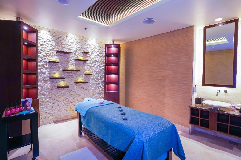 Mūsdienīgs spa telpa ar masāžas galdu, dekorēta ar akmens sienu un siltām gaismām, radot relaksējošu atmosfēru. Uz galda novietoti akmeņi un ziedi, pieejami masāžas eļļu pudelītes.