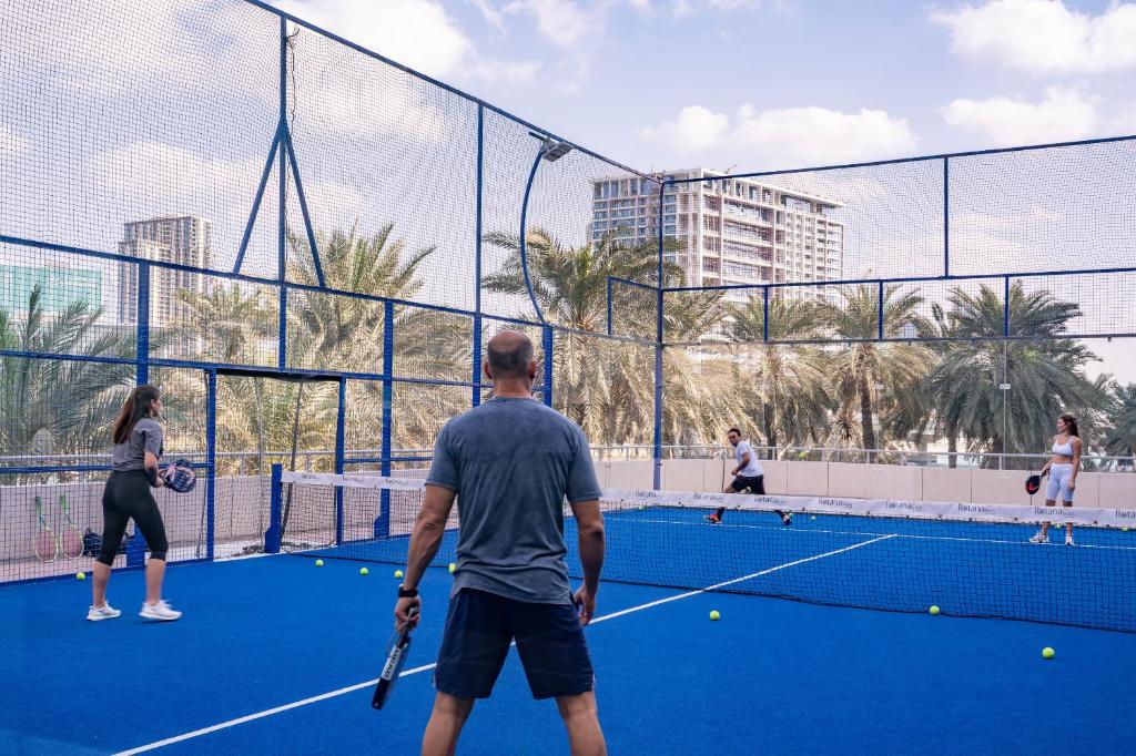 Tennisa spēle uz zilo laukumu, kurā piedalās četri spēlētāji, apkārt palmām un mūsdienīgām ēkām. Spēlētāji izmanto tennisa raketes un bumbiņas.
