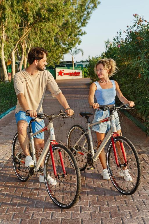 Jauna pāra smaidīšana un mijiedarbība, stāvot blakus saviem velosipēdiem, saulainā laikā. Fons ir apzaļumots ceļš, kas ved uz viesnīcu.