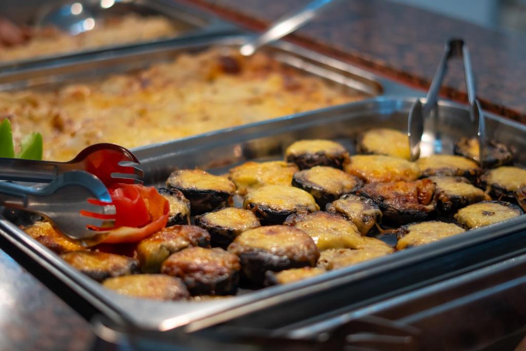 Ceptas baklažānu šķēles ar sieru un tomātiem, pasniegtas bufetē. Blakus redzama ceptu kartupeļu ēdiens.