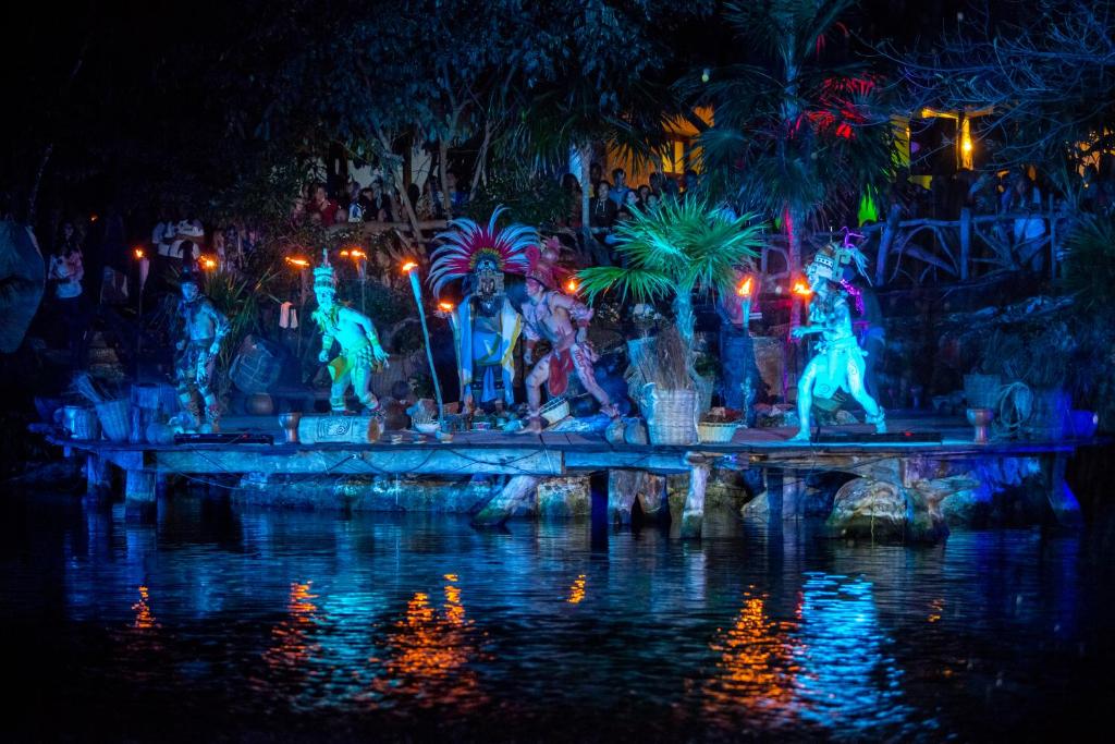 Nakts gaismas izrāde ar dejām un tradicionāliem tērpiem pie ūdens, apgaismota ar krāsainām gaismām un skatītājiem fona.