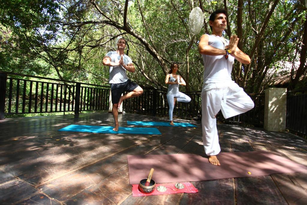 Jogas nodarbība dabā, kur trīs cilvēki praktizē dažādas pozas uz paklājiem, apzaļumotā vidē. Uz grīdas novietoti rīki meditācijai.