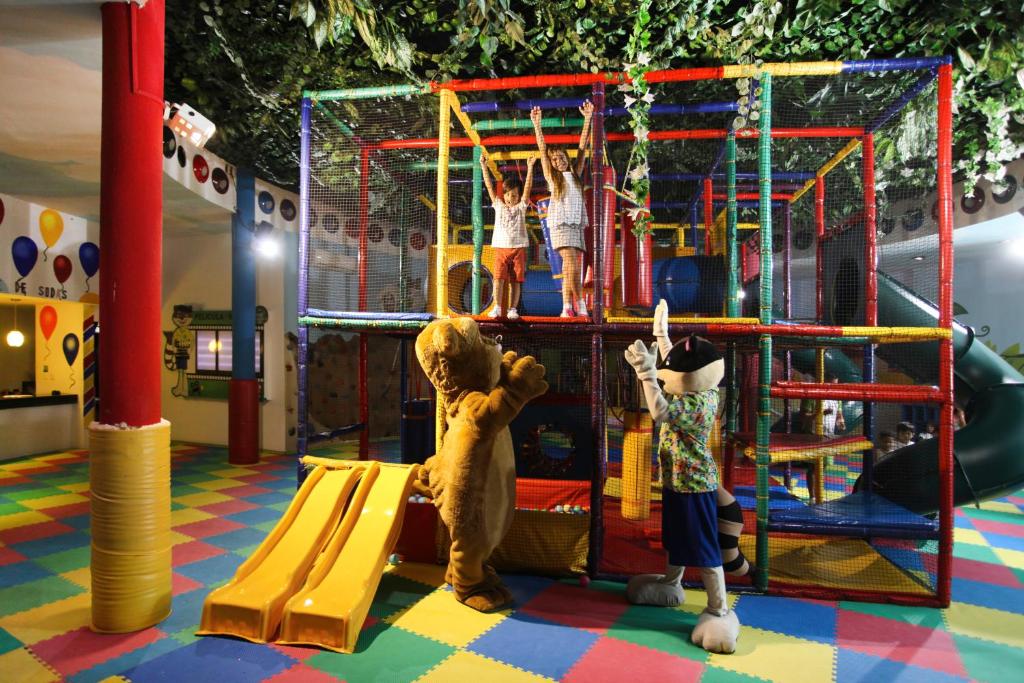 Bērnu rotaļu laukums ar krāsainu struktūru un slidkalniņu, kur bērni spēlējas un priecājas kopā ar maskotām figūrām. Rotaļu telpa ir noformēta ar dzīvnieku tēmu un krāsainām grīdām.
