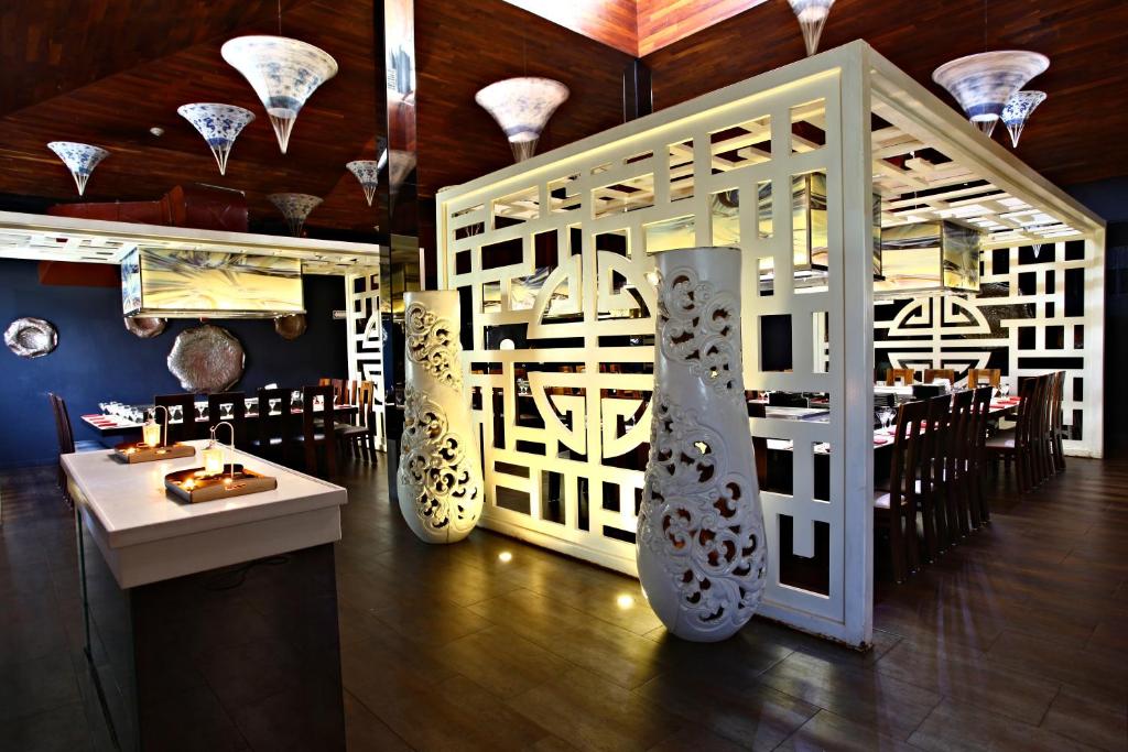 Mūsdienīgs restorāns ar elegantu dizainu, kurā redzama dekoratīva baltu paneļu struktūra un stilīgas gaismas. Galdi ir sakārtoti, gatavi viesu uzņemšanai.