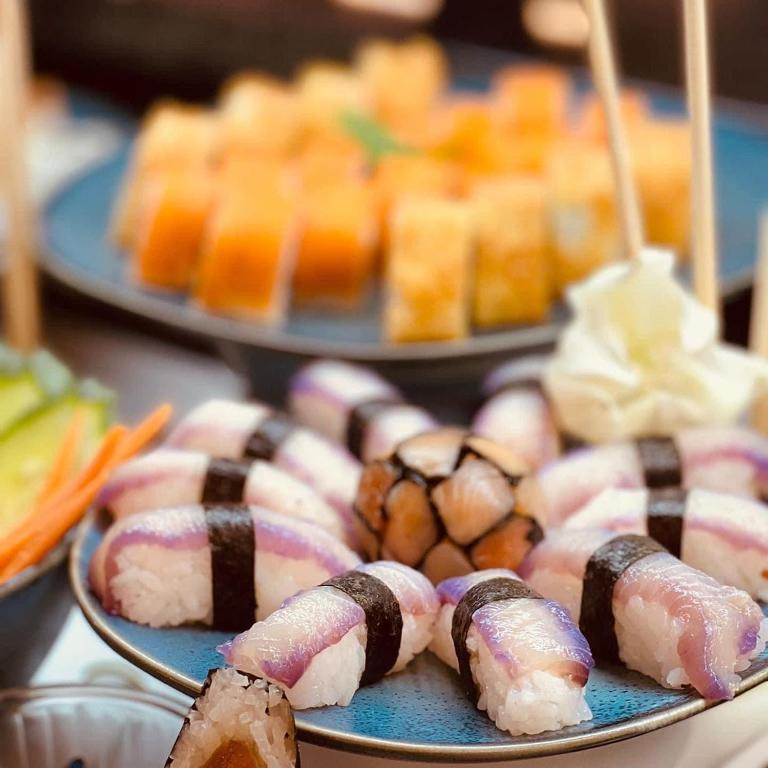 Skaisti pasniegts sushi šķīvis, kas satur dažādu veidu suši rullīšus un dārzeņu garnējumu. Fons ir citi suši un uzkodas.