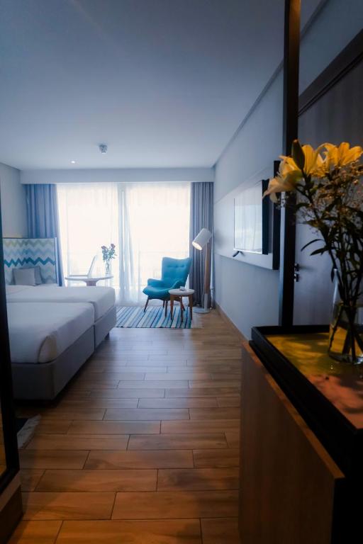 Mūsdienīgs viesnīcas numurs ar plašu logu, kas ļauj ieplūst dabiskai gaismai, un komfortablu zilu krēslu. Telpā redzami koka grīdas dēļi un elegants interjers, kas rada siltu atmosfēru.