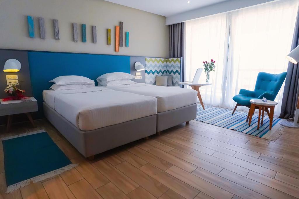 Mūsdienīgs viesnīcas numurs ar divām gultām, gaišām krāsām un stilīgu interjeru, kurā ir zils krēsls un galdiņš. Logi ļauj ieplūst dabīgai gaismai, radot patīkamu atmosfēru.