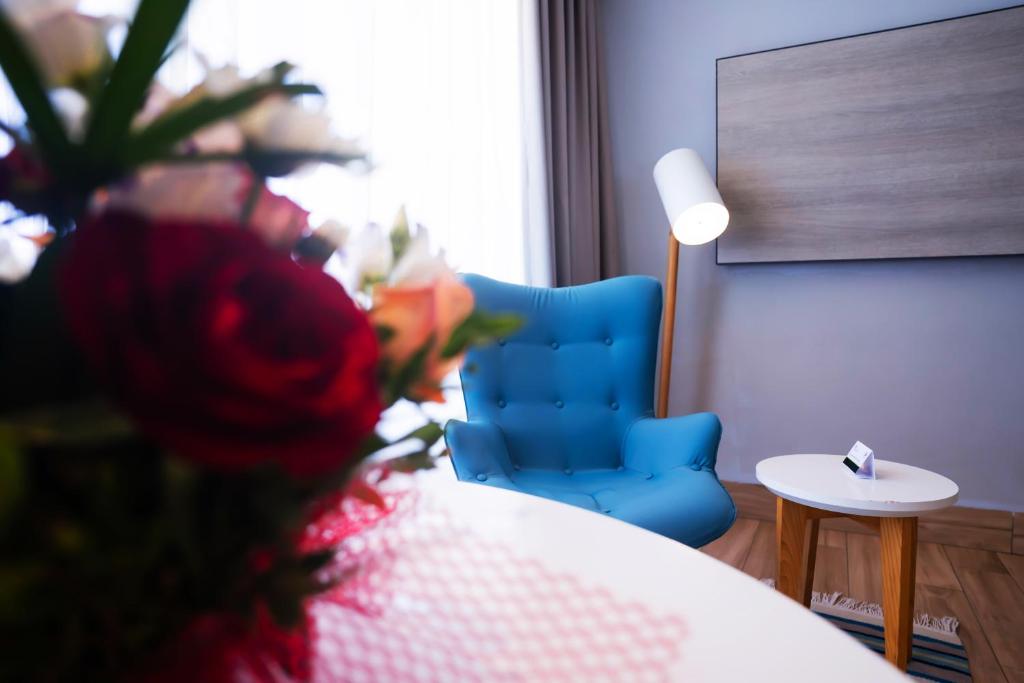 Mūsdienīgs viesistabas stūris ar zilu krēslu, balto galdiņu un ziedu pušķi priekšplānā. Gaisma ieplūst caur logu, radot siltu atmosfēru.