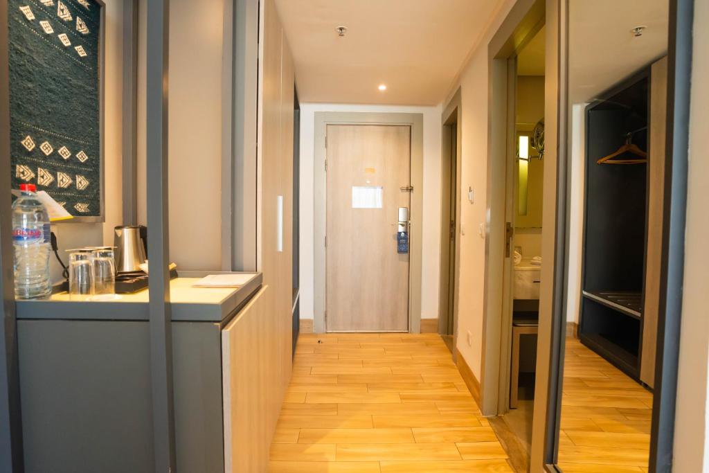 Mūsdienīgs viesnīcas numura gaiteņs ar koka grīdu, skatu uz durvīm un iekārtotu telpu. Redzama ūdens pudele, kafijas automāts un spogulis.