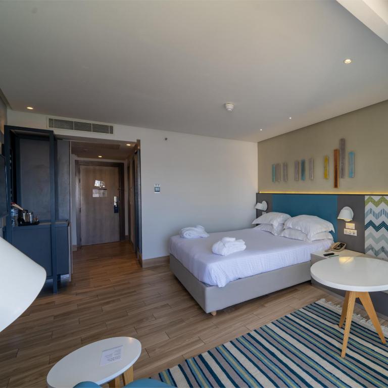 Mūsdienīga viesnīcas numura interjers ar divguļamo gultu, baltām dvieļiem un stilīgu dekora elementu kombināciju. Numurā ir arī kafijas automāts un neliela galdiņa zona.