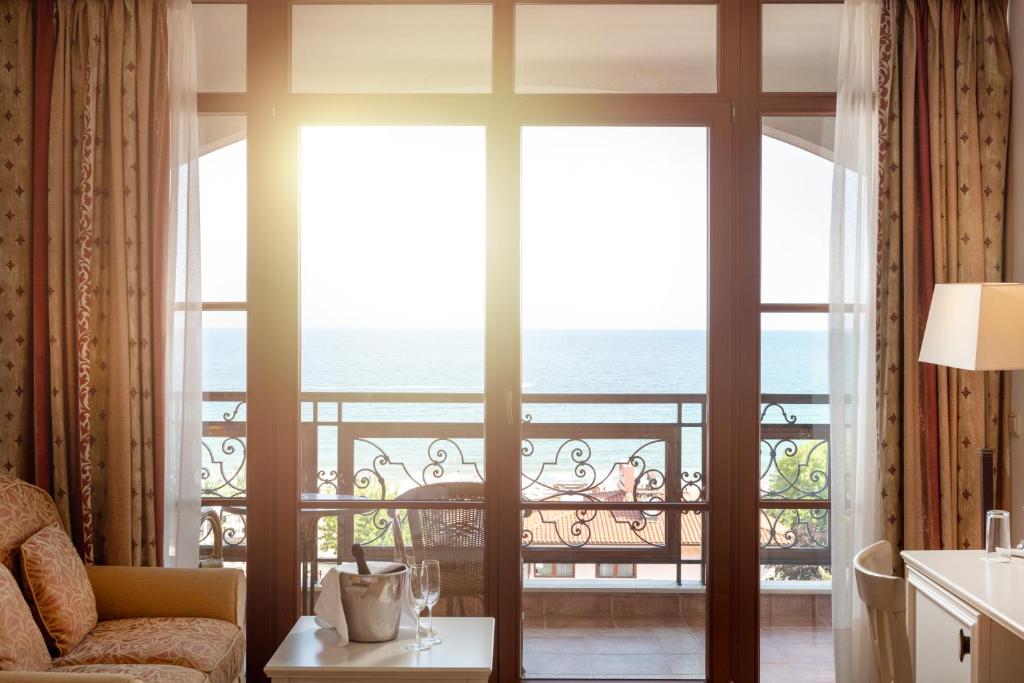 Apskate uz jūru no viesnīcas numura, kur saules gaisma ieplūst caur logiem, radot siltu un viesmīlīgu atmosfēru. Logā redzami balkona krēsli un galdiņš ar dzērieniem.