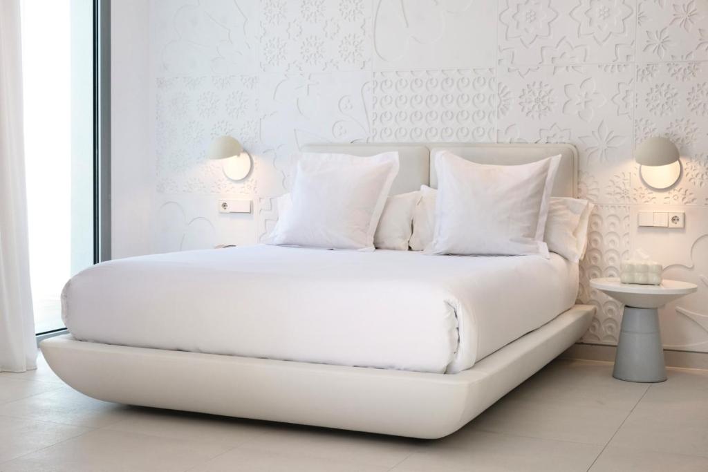 Mūsdienīga guļamistaba ar baltu gultu, mīkstiem spilveniem un minimālistisku dizainu, kas rada mierīgu un elegantu atmosfēru. Bērnu gultas galvgalis ir dekorēts ar reljefiem ziedu motīviem, bet blakus atrodas stilīgs naktsgaldiņš ar lampu.