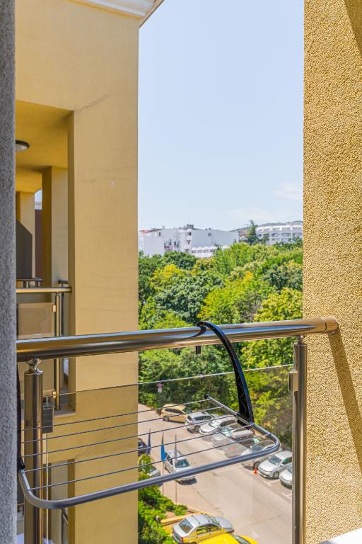 Skats no balkona uz zaļu apkaimi ar automašīnām uz ielas. Redzams balkona drēbju žāvētājs un gaišs, saulains laiks.