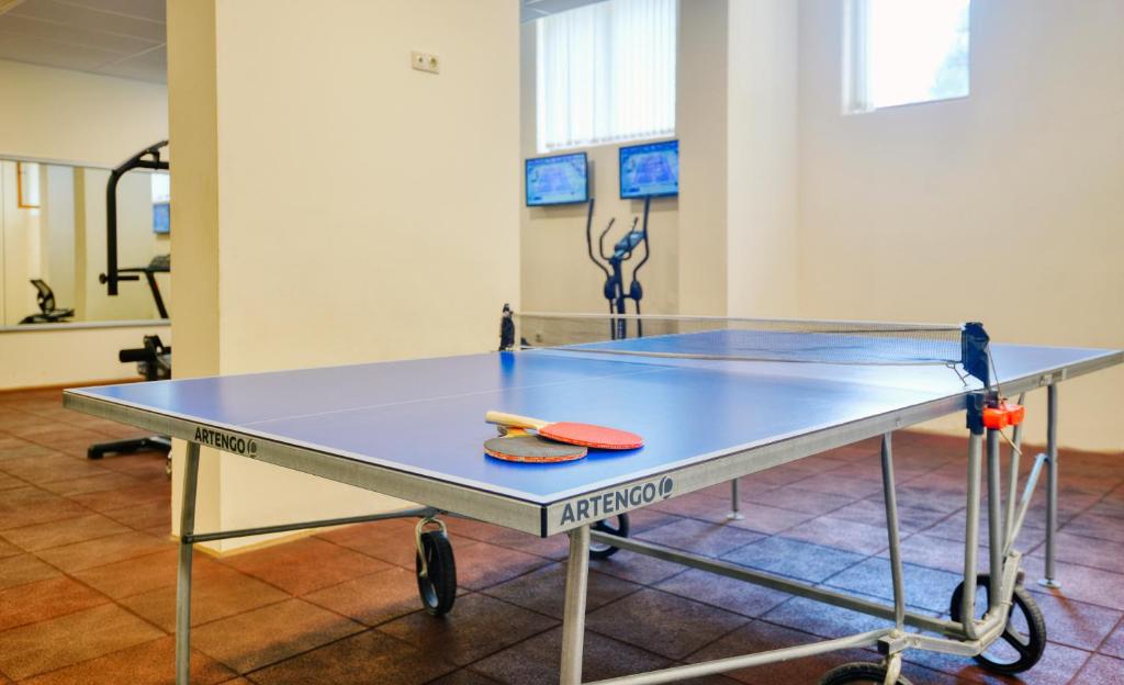 Pingponga galds ar raketēm un bumbiņu sporta zālē, fona ekrānos redzami sporta pasākumi.