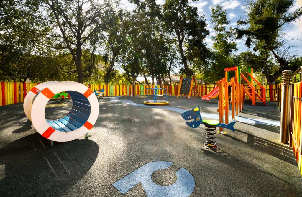 Rotaļu laukums ar krāsainām spēļu iekārtām, including slidkalniņu, šūpolēm un rotaļu tuneli. Apkārt laukuma ir ziedošas koku lapas un daudzkrāsaini žogi, radot jautru un draudzīgu atmosfēru.