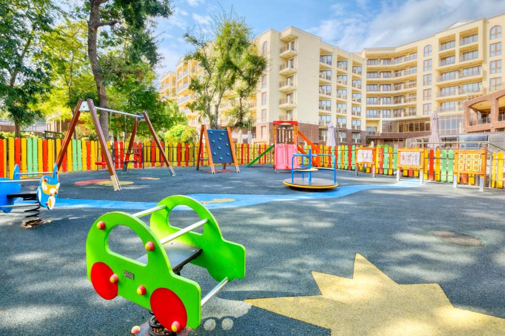 Krāsains bērnu laukums ar šūpolēm, kāpšanas sienu un rotaļu elementiem, apkārt koša žoga un daudzdzīvokļu ēkas fonā. Ideāla vieta bērnu rotaļām un izklaidei.