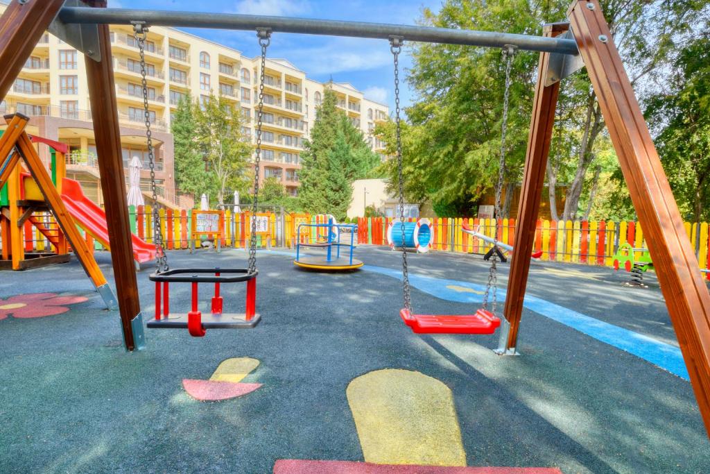 Krāsains bērnu rotaļu laukums ar šūpolēm, slidkalniņu un griežamo platformu, apjozts ar krāsainu žogu un kokiem. Ideāla vide bērnu spēlēm un izklaidei.