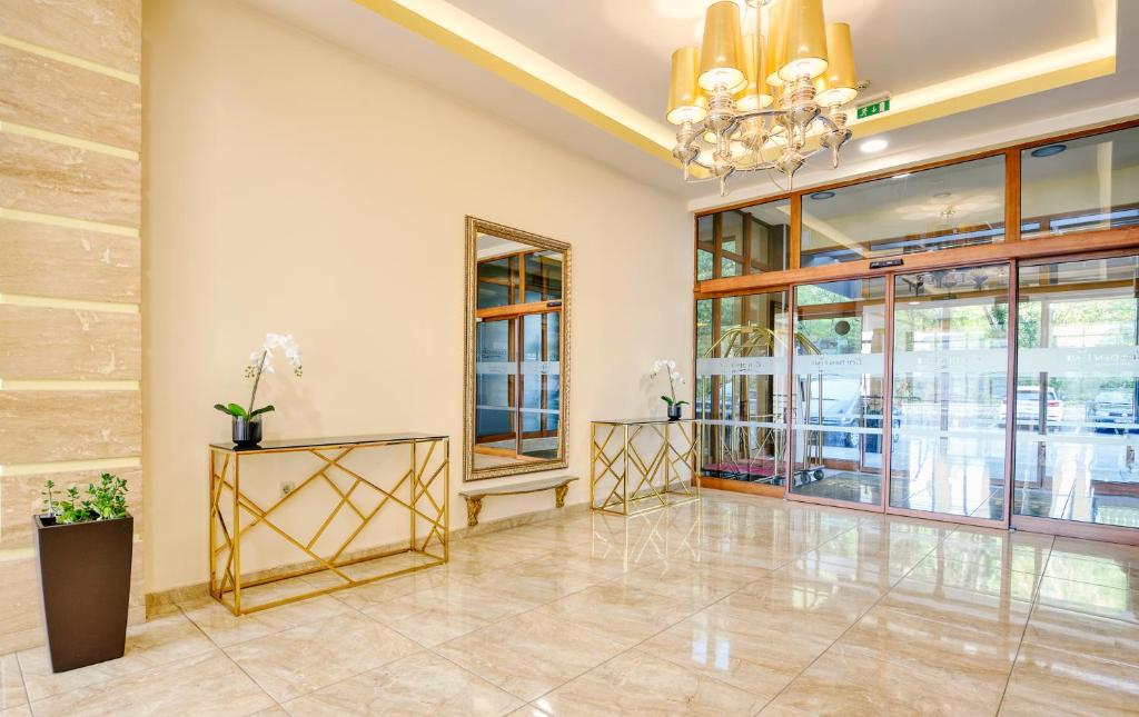 Grezna viesnīcas uzgaidāmā telpa ar mirdzošām flīzēm, elegantu griestu lampu un izsmalcinātiem zelta toņu mēbelēm. Telpā ir dabiski elementi, piemēram, orhidejas un zaļie augi.