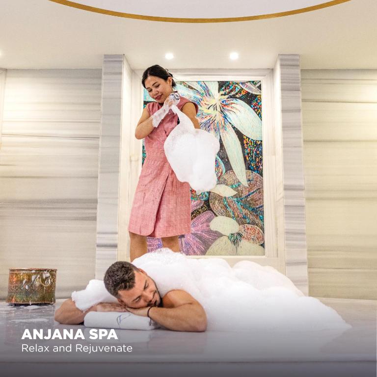 Spa procedūra ar putām, kurā sieviete apkalpo vīrieti, kas guļ uz galda. Fons rotāts ar krāsainu mākslas darbu.