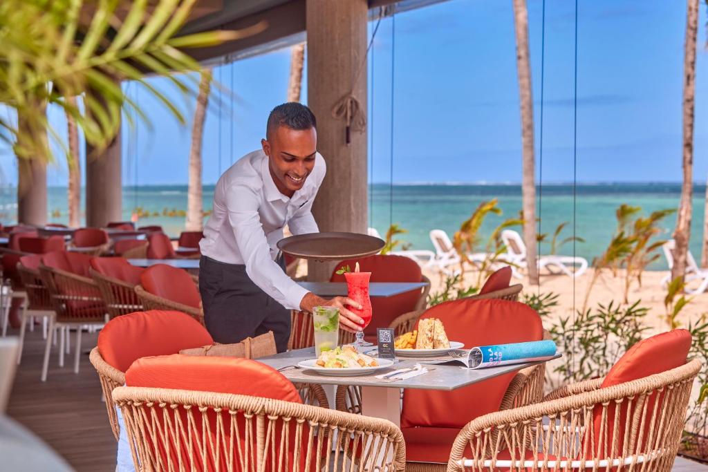 Viesmīlis apkalpo viesus pludmales restorānā, pasniedzot dzērienus un ēdienus pie galda ar skatu uz jūru.