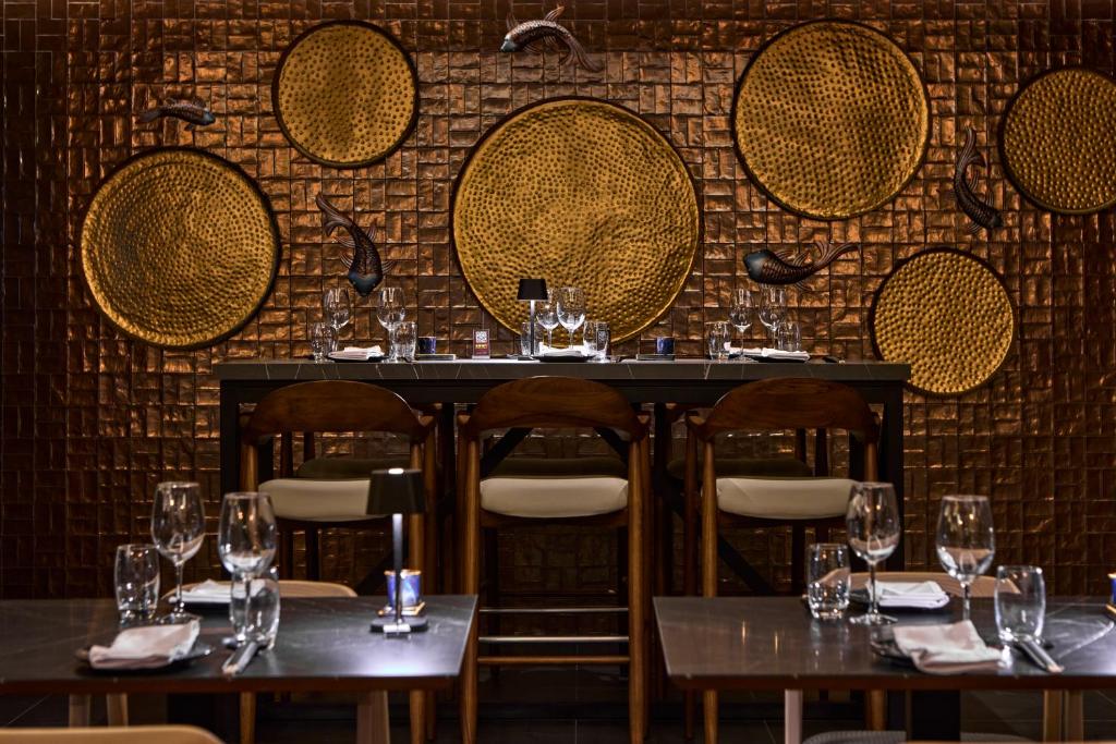 Eleganta restorāna interjers ar metāla dekoriem un galdiem, kas sagatavoti viesu uzņemšanai. Sienas rotā izsmalcināti apļa formas mākslas elementi.