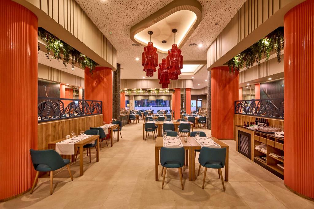 Mūsdienīgs restorāna interjers ar krāsainiem stabiem un elegantiem galdiem, kas izveidots patīkamai ēšanas atmosfērai. Izceltas sarkanas laternas un zaļie augi piešķir dzīvīgumu telpai.