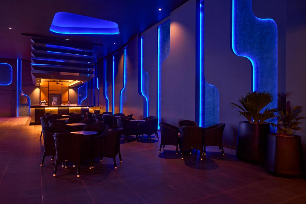 Mūsdienīga bāra iekārtojums ar zilu apgaismojumu un elegantiem krēsliem, kas radīts atpūtas atmosfērai. Interjers izceļas ar stilīgu dizainu un augu akcentiem.