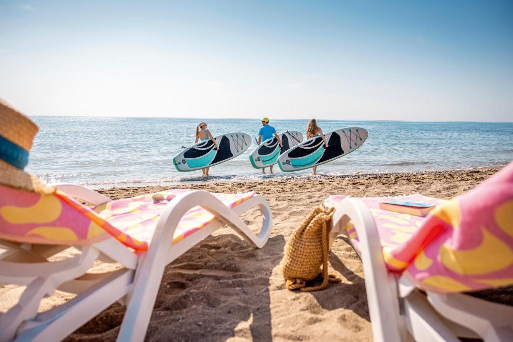 Trīs cilvēki ar paddleboardiem dodas uz jūru, baudot saulainu pludmali un mierīgu ūdeni. Priekša redzami atpūtas krēsli un pludmales aksesuāri.