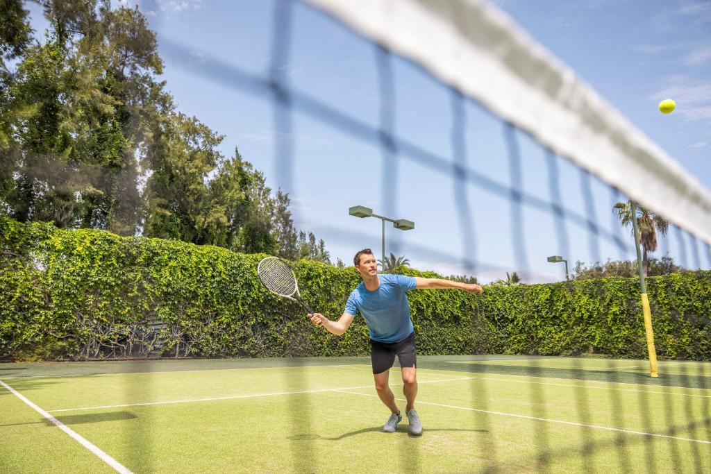 Vīrietis spēlē tenisu, sitot bumbu uz laukuma ar raketi. Fons ir zaļš, ar kokiem un lampām, kas norāda uz saulainu dienu.