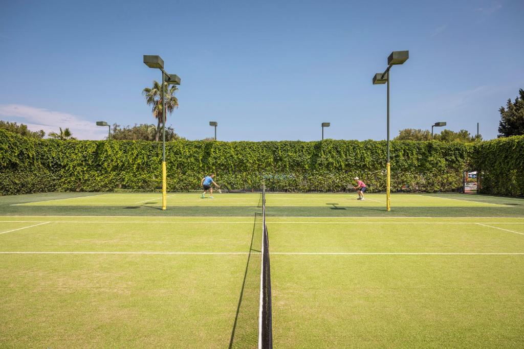 Tenisa laukums ar diviem spēlētājiem, kas spēlē pretējiem galiem, zaļā zālē un apstādījumos. Gaismas stabi nodrošina apgaismojumu, bet debesīs ir skaidrs laiks.