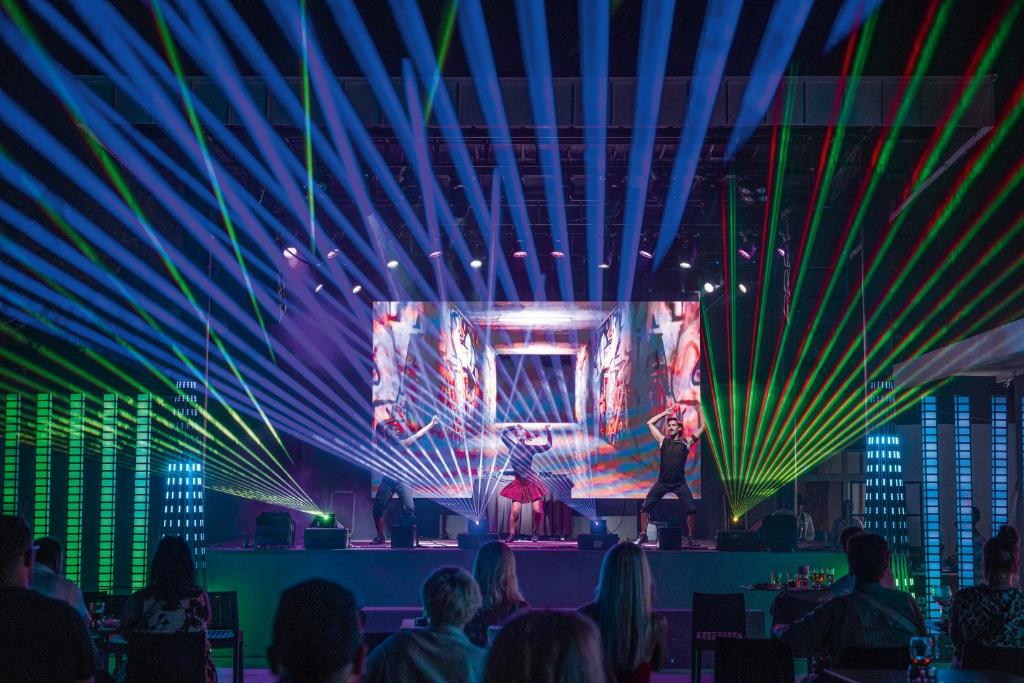 Skatuves priekšnesums ar dejotājiem un krāsainiem gaismas stariem, radot dinamisku atmosfēru. Aiz dejotājiem ir liels ekrāns ar vizuālo materiālu.