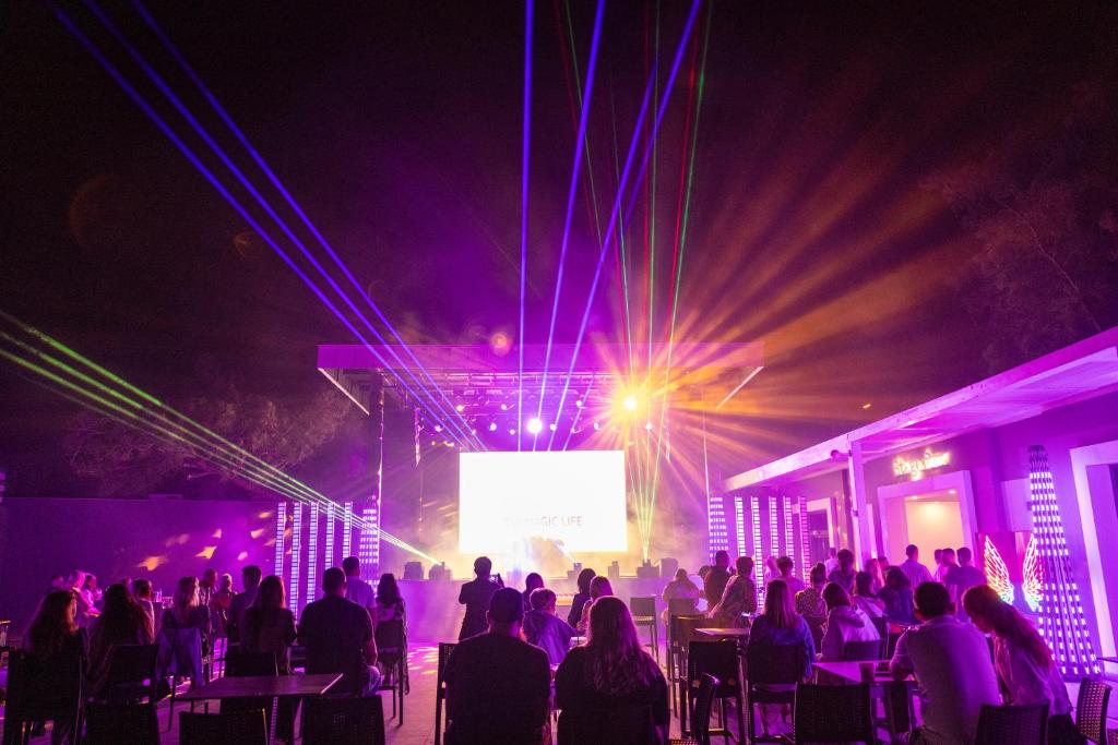 Nakts pasākums ar krāsainiem gaismas stariem un skatītājiem, kas vēro izrādi uz lielā ekrāna. Atmosfēra ir dzīvīga un enerģiska.